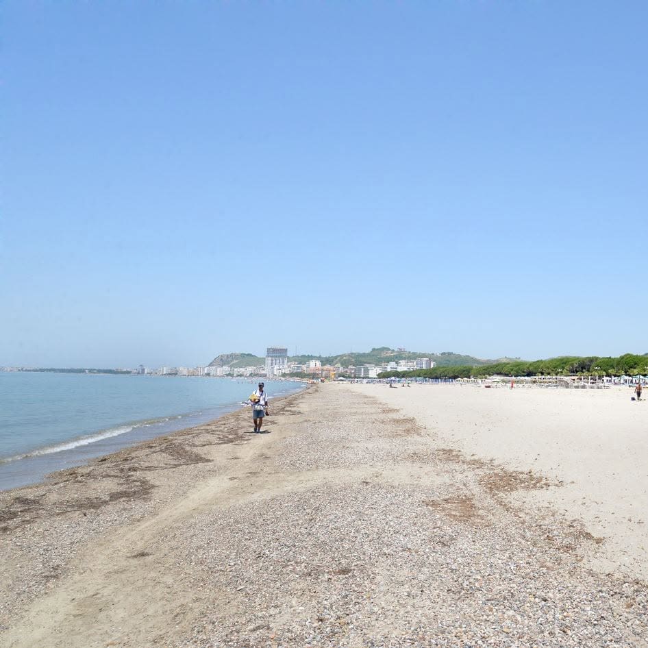 A long sandy beach beside calm sea, with a man walking at the waterline, sunshades under pine trees and a town on distant hills