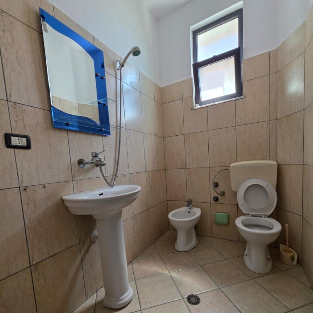 A tiled bathroom with a pedestal basin under a mirror, a hand shower on the wall above it, a bidet, a toilet and a floor drain