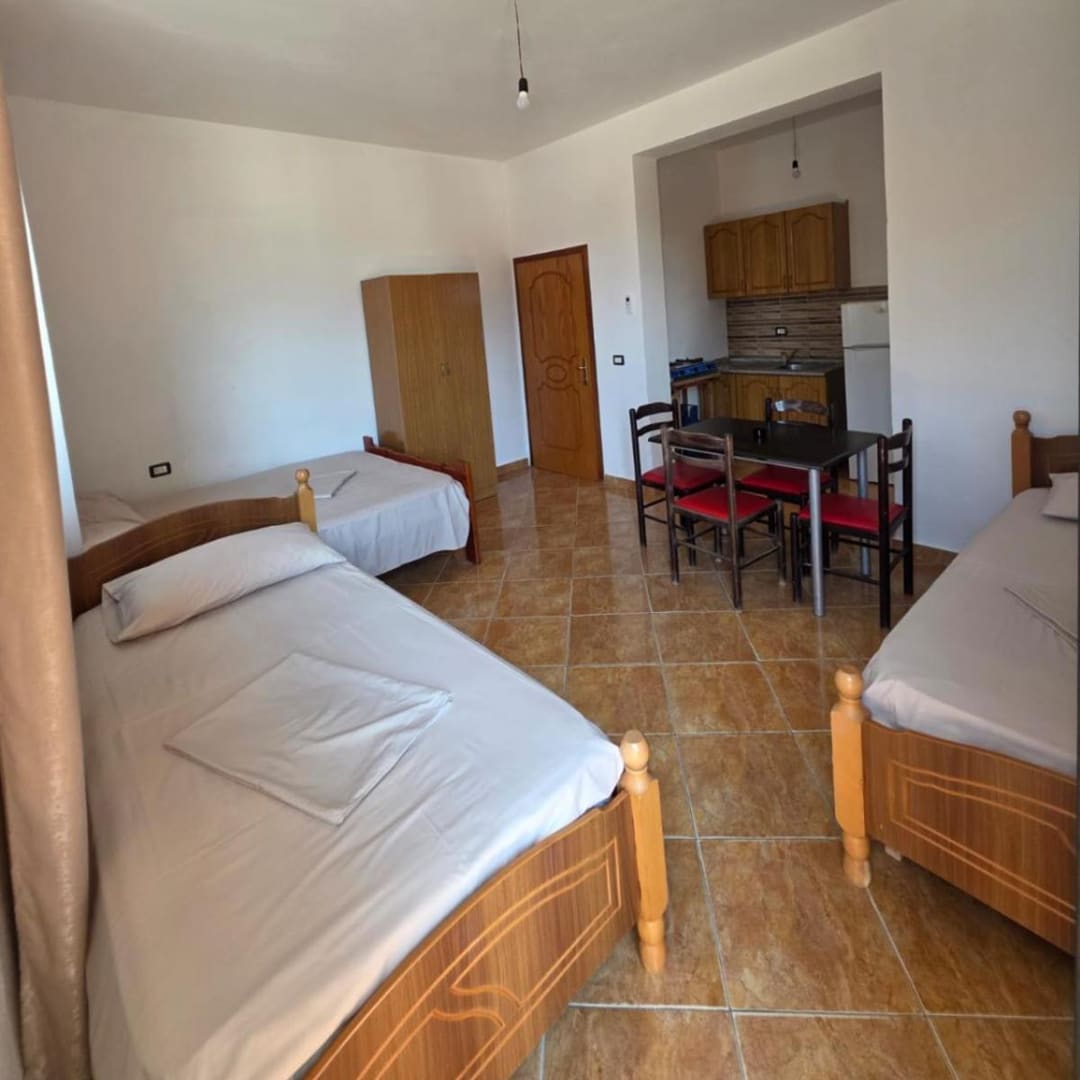 Three beds, a wardrobe and a table with red chairs in a tiled room, with a kitchenette and a fridge in an alcove behind