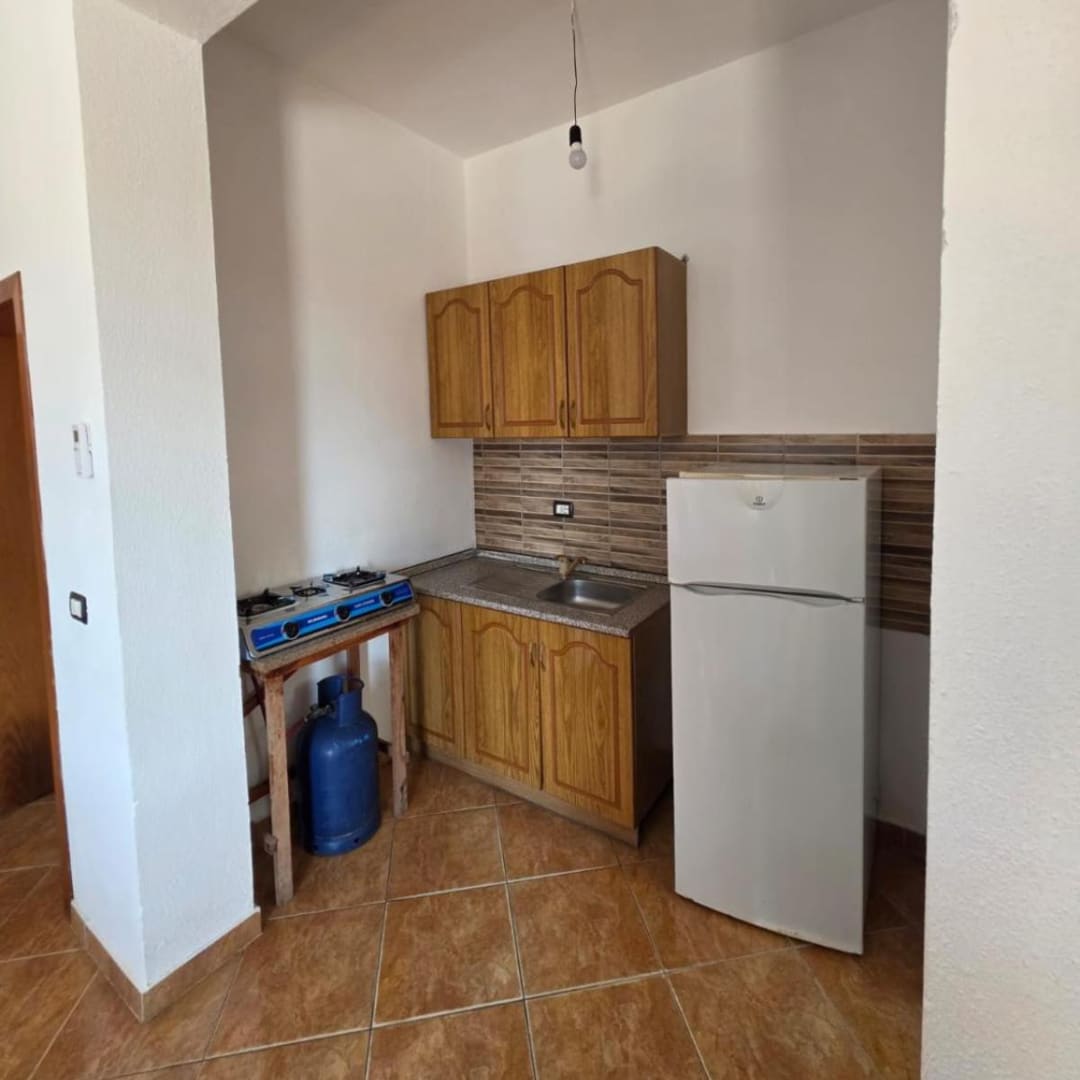 A kitchenette in an alcove with wooden cupboards, a sink, a gas hob on a wooden stand above a gas bottle, and a white fridge