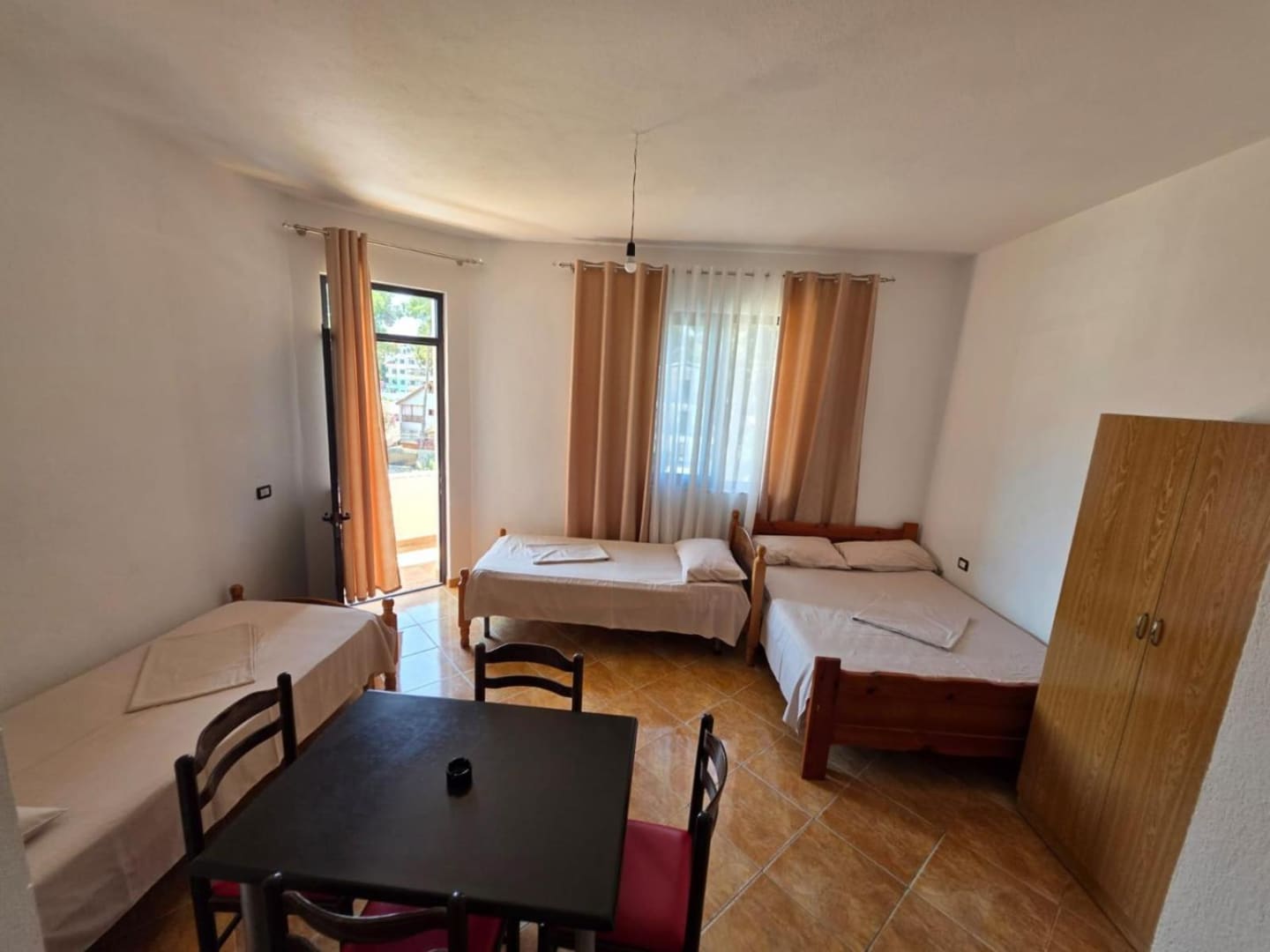 Three beds, a dining table and a wardrobe in a tiled room, with the balcony door open